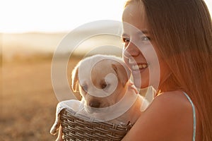Happy girl holding her little puppy dog in a basket looking at the sun setting - close up
