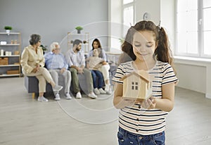 Happy girl holding toy house standing in new apartment with her family in background