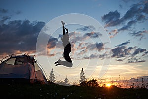 Happy girl jumping on grass with wildflowers at camping in mountains at dawn under blue sky