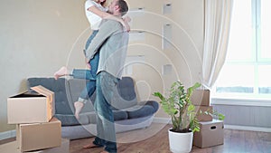 A happy girl jumps on the back of her cheerful guy in a new apartment among the boxes. Young couple moving in a new home