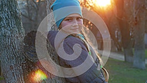 Happy girl with long braided hair portrait in spring park sits on a tree on sunset outdoor. Delighted little kid in trendy coat an