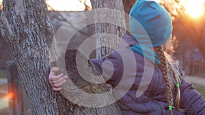 Happy girl with long braided hair portrait in spring park sits on a tree on sunset outdoor. Delighted little kid in trendy coat an