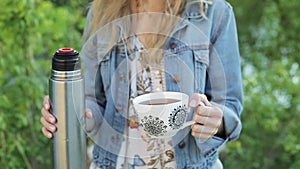 Happy girl with long hair holds a cup of tea or coffee in hands against the blurred nature background. Pretty woman in