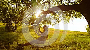 Happy girl with long hair, swinging on swing on tree branch, in bright rays of a golden sunset and smiling. Slow motion.