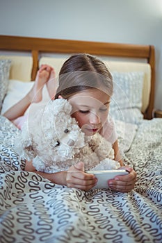 Happy girl lying on bed with a teddy bear and using mobile phone