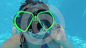 Happy girl in a mask under the water in the swimming pool looks into the camera, bubbles