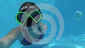 Happy girl in a mask under the water in the swimming pool looks into the camera, bubbles