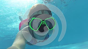 Happy girl in a mask under the water in the swimming pool looks into the camera, bubbles, sun rays