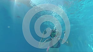 Happy girl in a mask under the water in the swimming pool looks into the camera, bubbles, sun rays