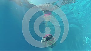 Happy girl in a mask under the water in the swimming pool looks into the camera, bubbles, sun rays