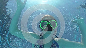 Happy girl in a mask under the water in the swimming pool looks into the camera, bubbles, sun rays