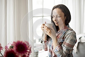 Happy girl with a mug of fragrant coffee in hands. Good breakfast at home in the living room by the window. Free space for text.