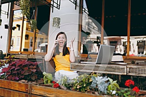 Happy girl in outdoors street cafe sitting at table with laptop pc computer, talking on mobile phone, conducting