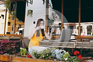 Happy girl in outdoors street coffee shop cafe sitting at table with laptop pc computer, texting message on mobile phone