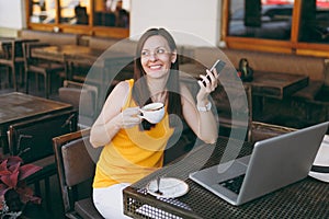 Happy girl in outdoors street coffee shop cafe sitting at table with laptop pc computer, texting message on mobile phone