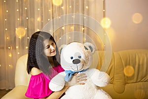 Happy girl in a pink dress hugging a big white soft bear home on the couch. Gift from a loved one, valentine`s day, birthday,