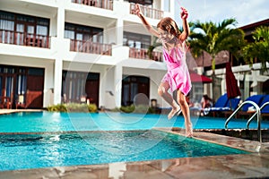 Happy girl in pink swimsuit jumps with joy into outdoor pool at sunny resort. Child enjoys summer vacation, fun