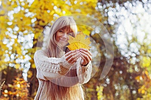 Happy girl playing with leaf, looking at camera and smilling. Autumn portrait woman hides her face yellow maple leaf