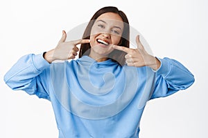 Happy girl pointing fingers at white perfect smile, straight whitened teeth, standing in hoodie against white background