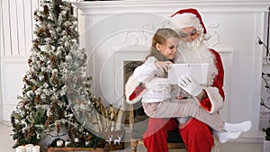 Happy girl and Santa Claus looking at presents on tablet PC next to the Christmas tree