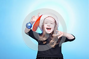 Happy girl in santa hat with christmas toys on blue background. Funny young child waiting for christmas, new year and presents.