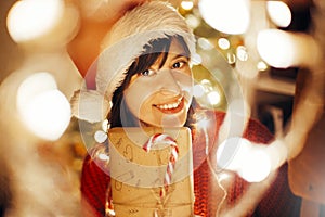 happy girl in santa hat and red sweater wrapping christmas presents in lights in evening festive room at tree illumination.