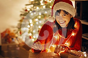 happy girl in santa hat and red sweater wrapping christmas presents in lights in evening festive room at tree illumination.
