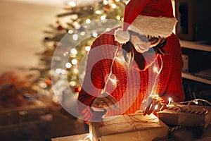 happy girl in santa hat and red sweater wrapping christmas presents in lights in evening festive room at tree illumination.