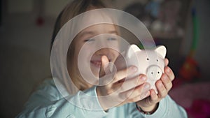 Happy girl save money in piggy bank in her home. Child inserting a coin into a piggy bank, indoor financial concept. Kid