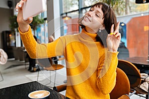 Happy girl showing peace sign and taking selfie on cellphone in cafe