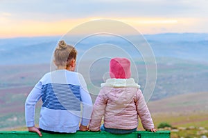 Happy girl sister friends sitting on bench at beautiful landscape background spring field and sunset sky nature outdoors