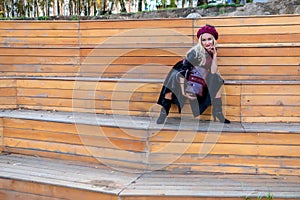A happy girl sits on a summer theater bench, made of wood, in a burgundy coat and biret, an adult with tender lips, in