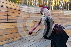 A happy girl sits on a summer theater bench, wooden in a burgundy coat and biret, smiles at the camera, in the fall