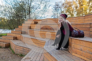 A happy girl sits on a wooden summer theater bench in a burgundy coat and biret, looking at the camera, in the fall