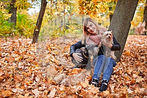 A happy girl sits in yellow autumn leaves in park and takes off her two little dogs.