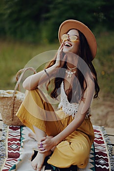 A happy girl is sitting in eco clothes on a plaid by the lake wearing a hat in a hippie look and smiling in surprise