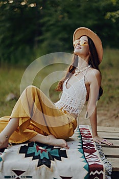 A happy girl sitting in eco clothing in nature on a plaid by the lake wearing a hat in a hippie look and smiling in