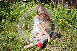 Happy girl sitting on the grass with a red cup of coffee on a summer morning.