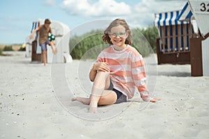 Happy girl sitting on white beach and brother on backround playing with ball at summer holidays