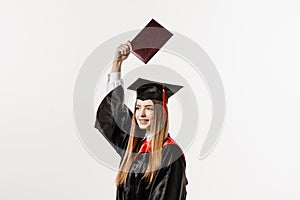 Happy girl student in black graduation gown and cap raises masters degree diploma above head on white background