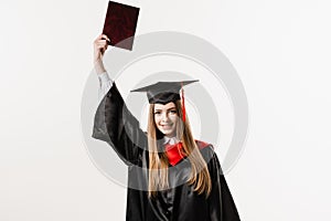 Happy girl student in black graduation gown and cap raises masters degree diploma above head on white background