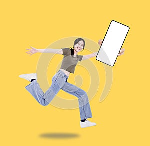 Happy girl student showing the blank screen mobile phone jump and run wear t-shirt denim isolated yellow background