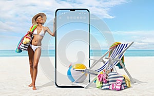 Happy girl in summer beach holiday wearing bikini showing a big screen mobile phone with deckchair and sea accessories isolated in