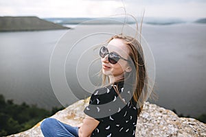 Happy girl in sunglasses sitting on top of rock mountain with beautiful view on river. Young tourist woman with windy hair