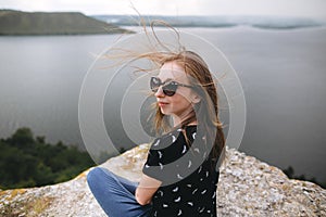 Happy girl in sunglasses sitting on top of rock mountain with beautiful view on river. Young tourist woman with windy hair