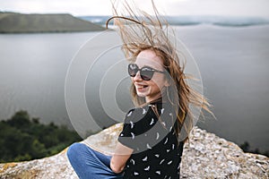 Happy girl in sunglasses sitting on top of rock mountain with beautiful view on river. Young tourist woman with windy hair