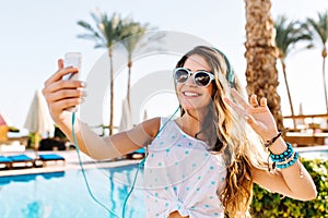 Happy girl in sunglasses with tanned skin making selfie with peace sign on palm trees background. Charming long-haired