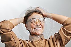 Happy girl touching head. Overjoyed caucasian woman with short haircut in glasses and beige jacket smiling. Portrait on