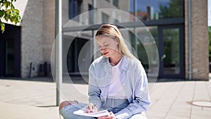 Happy girl university student writing in notebook studying sitting outdoor.