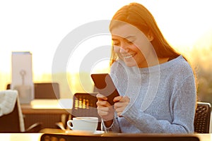 Happy girl using smartphone at sunset in a coffee shop